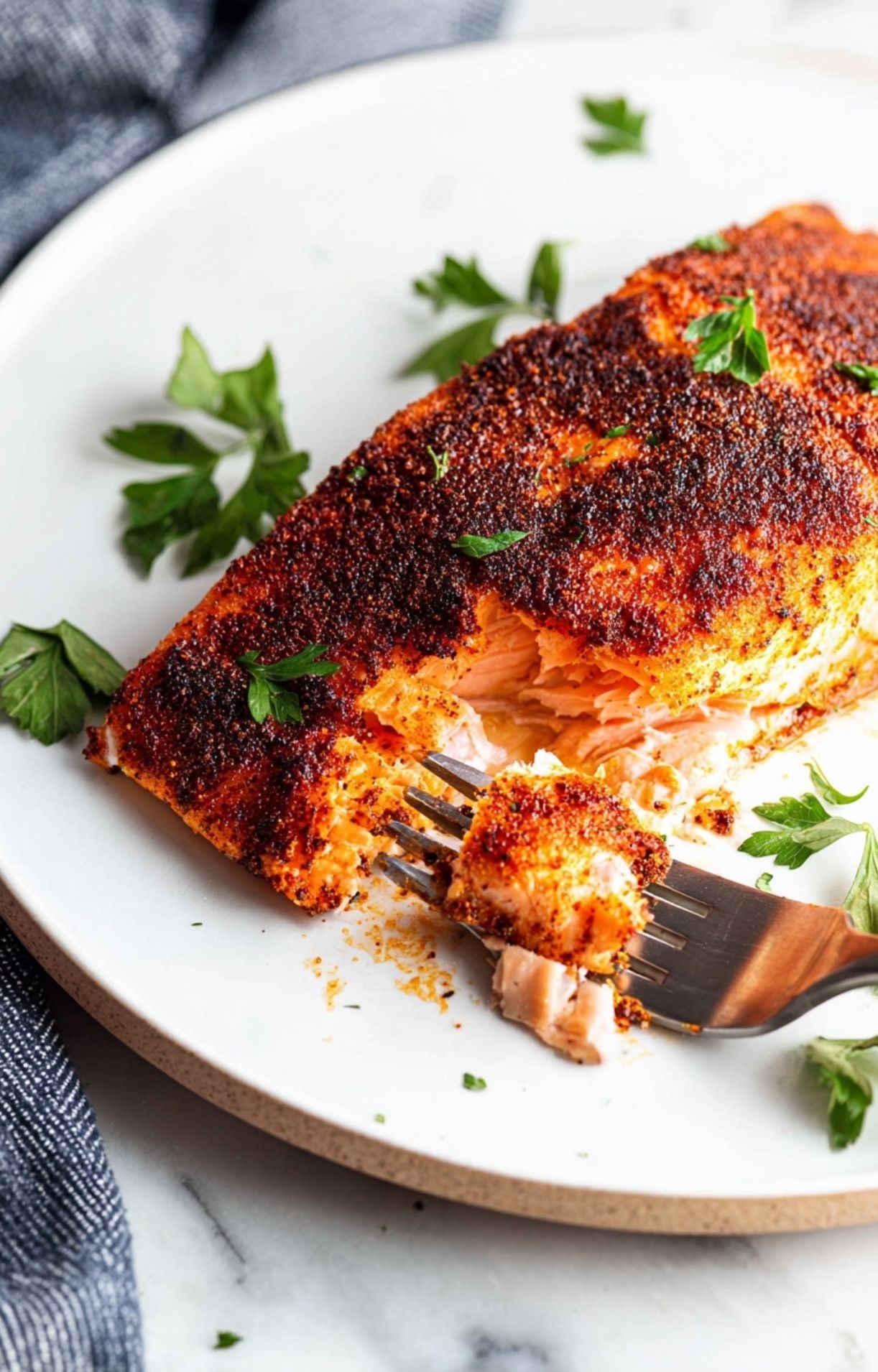 A final presentation shot of a juicy blackened salmon fillet on a white plate with lemon and parsley, with a fork holding up a perfect piece of the fish.