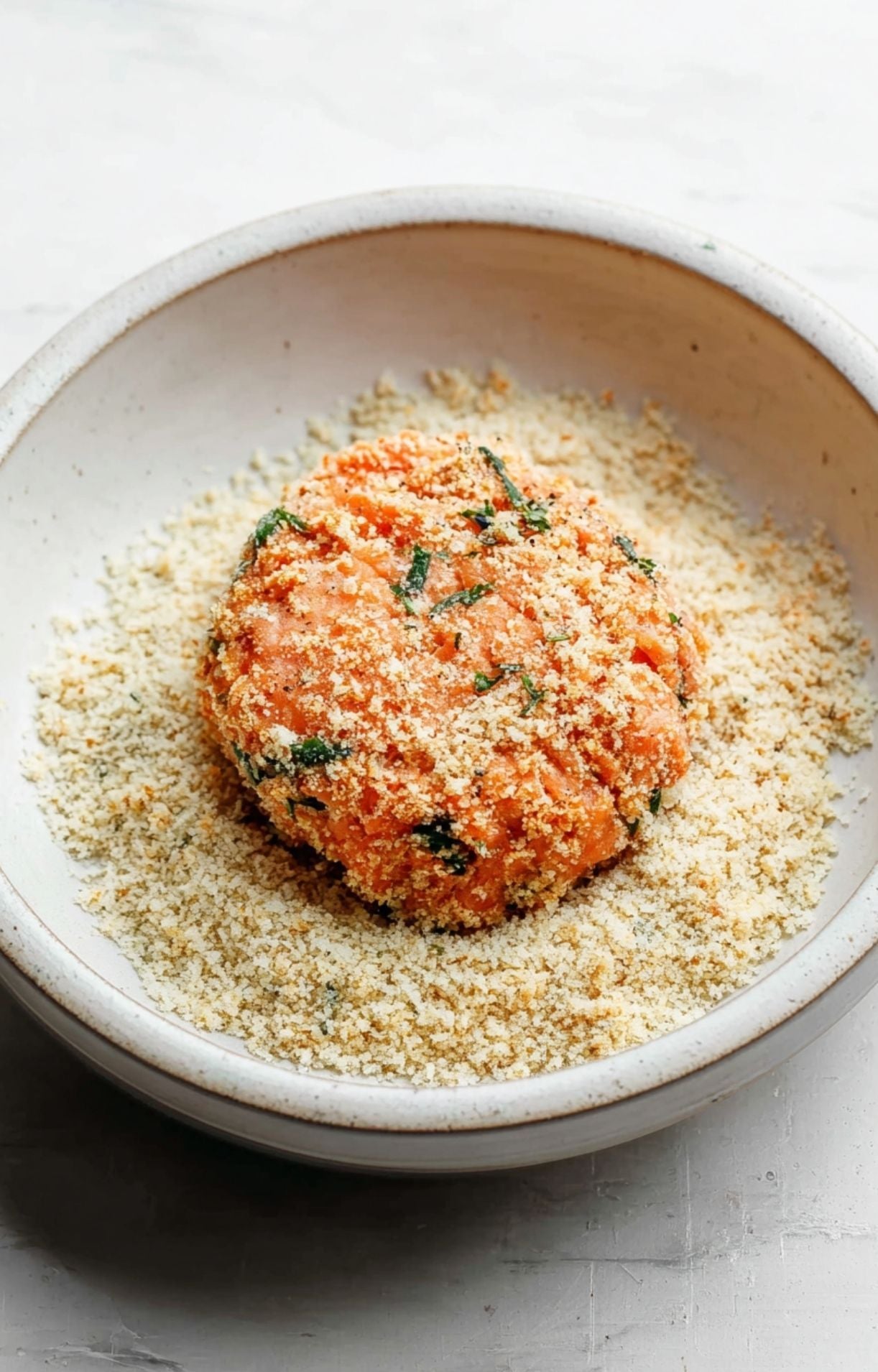A thick, raw salmon patty being pressed into a bowl of seasoned breadcrumbs for a crispy exterior.