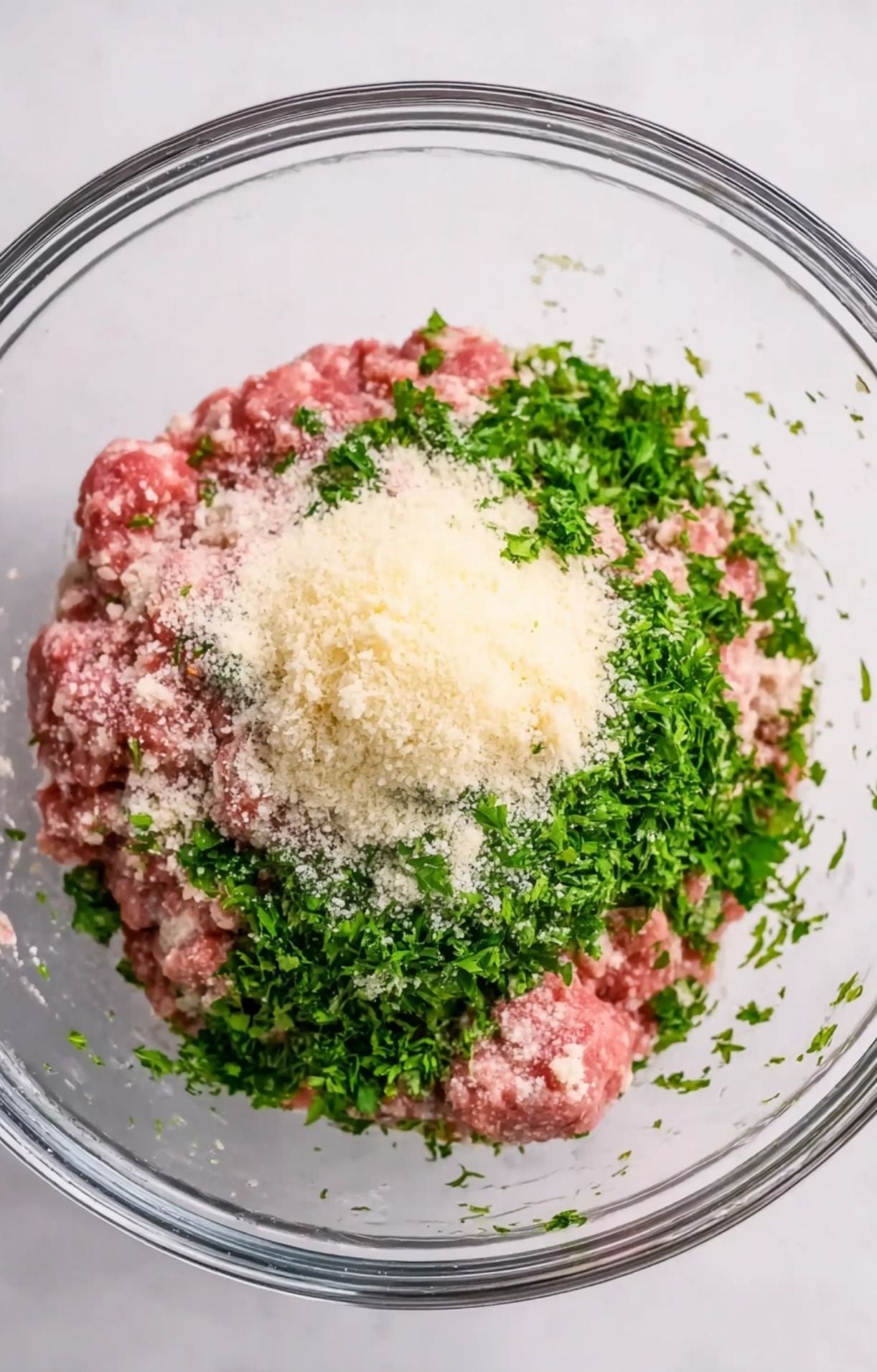A glass bowl containing ground meat topped with a large mound of freshly chopped bright green parsley and a pile of grated parmesan cheese.