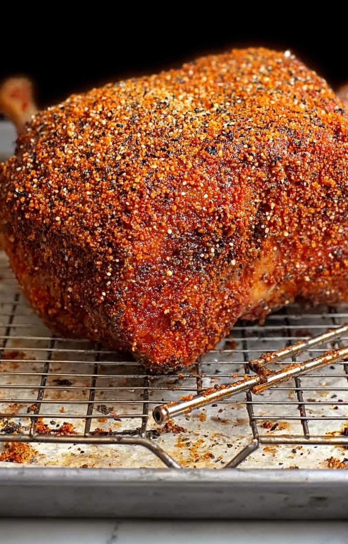 A raw prime rib roast completely coated in a thick layer of red and black spice rub, sitting on a wire rack over a baking sheet.