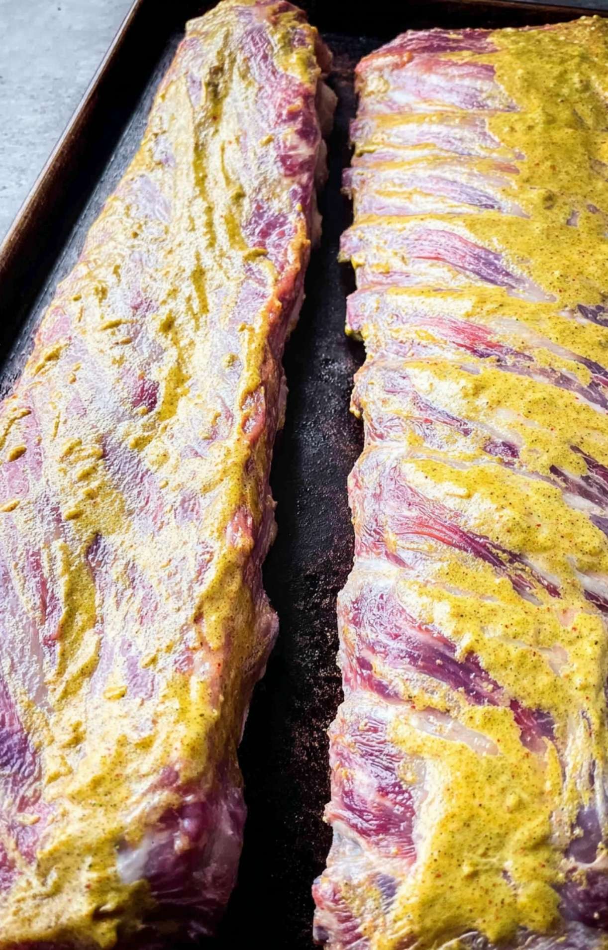 Two racks of baby back ribs being coated in yellow mustard binder on a baking sheet before smoking.