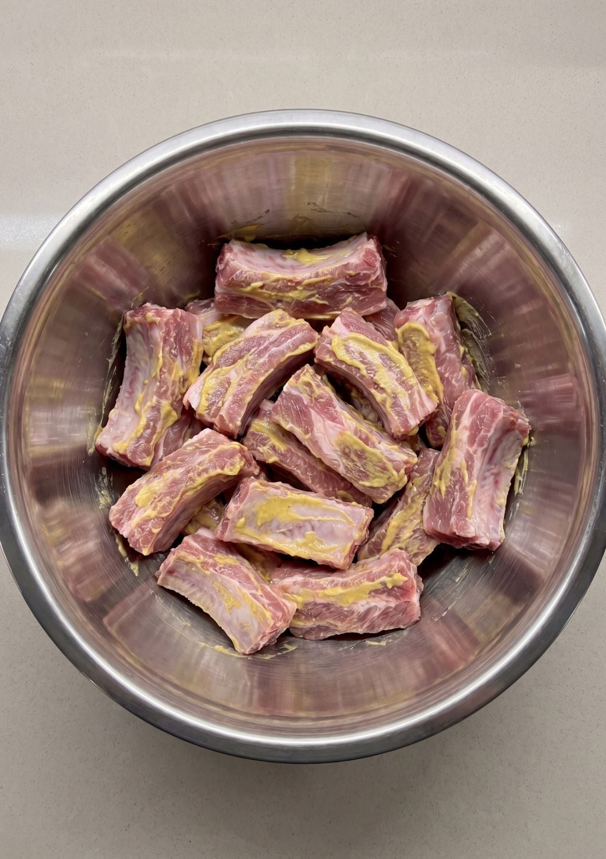 Raw individual rib pieces sitting in a large stainless steel mixing bowl coated in yellow mustard.
