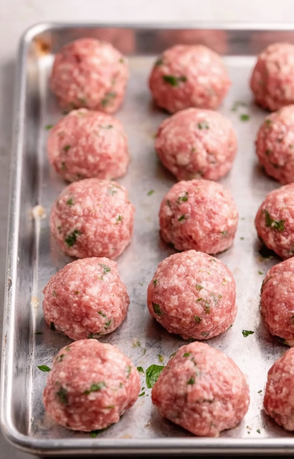 Rows of raw, hand-rolled meatballs flecked with green parsley arranged on a silver baking sheet before cooking.