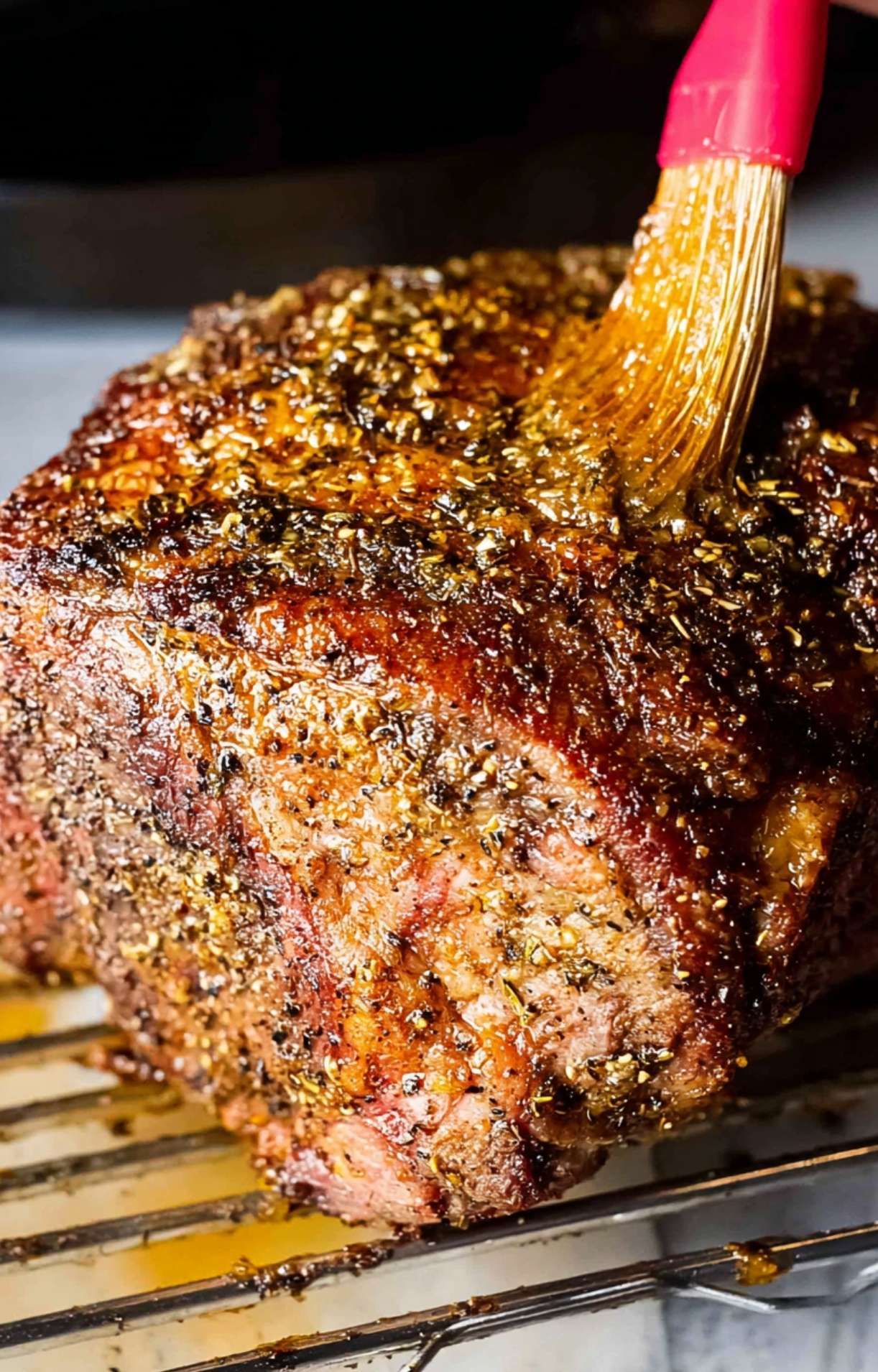 A silicone brush applying a finishing glaze or butter to the dark, seared crust of the prime rib during the smoking process.