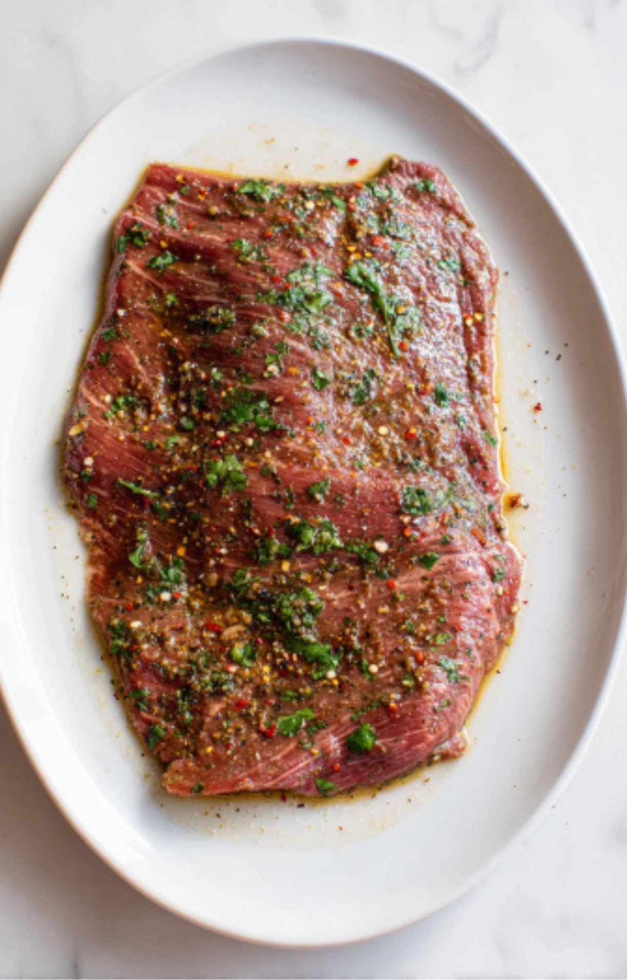 A raw flank steak coated in a herb and spice marinade on a white platter for a Flank Steak Salad Recipe.