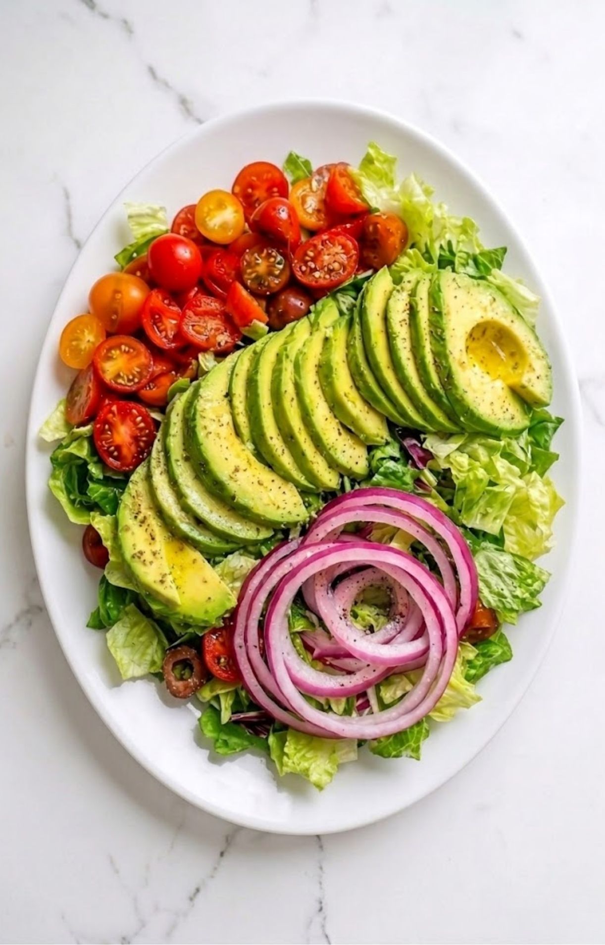 A white plate topped with a bed of green lettuce, neatly arranged sliced avocado, cherry tomato halves, and red onion rings.