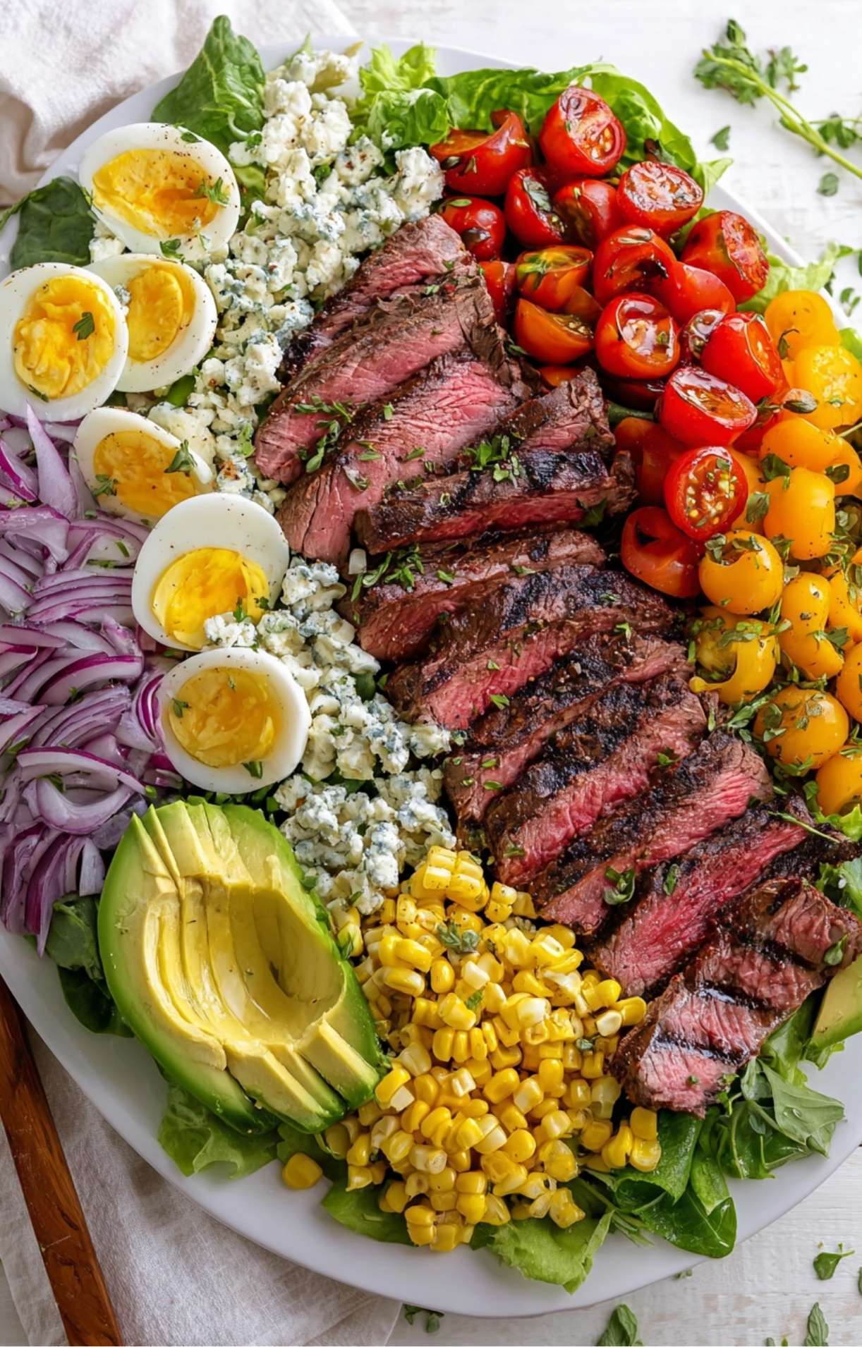 A high-angle view of a densely packed salad platter featuring medium-rare steak as the centerpiece surrounded by classic Cobb ingredients.