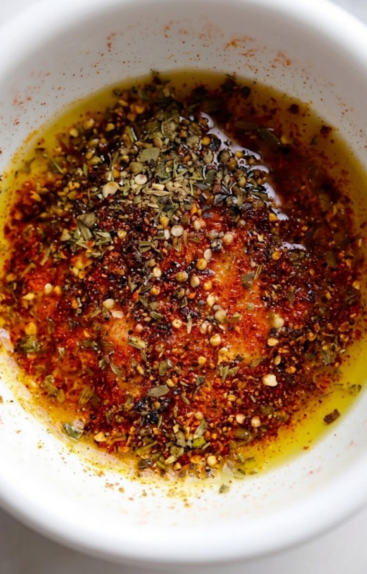 A white bowl containing a mixture of olive oil and dried spices including paprika, oregano, and red pepper flakes.