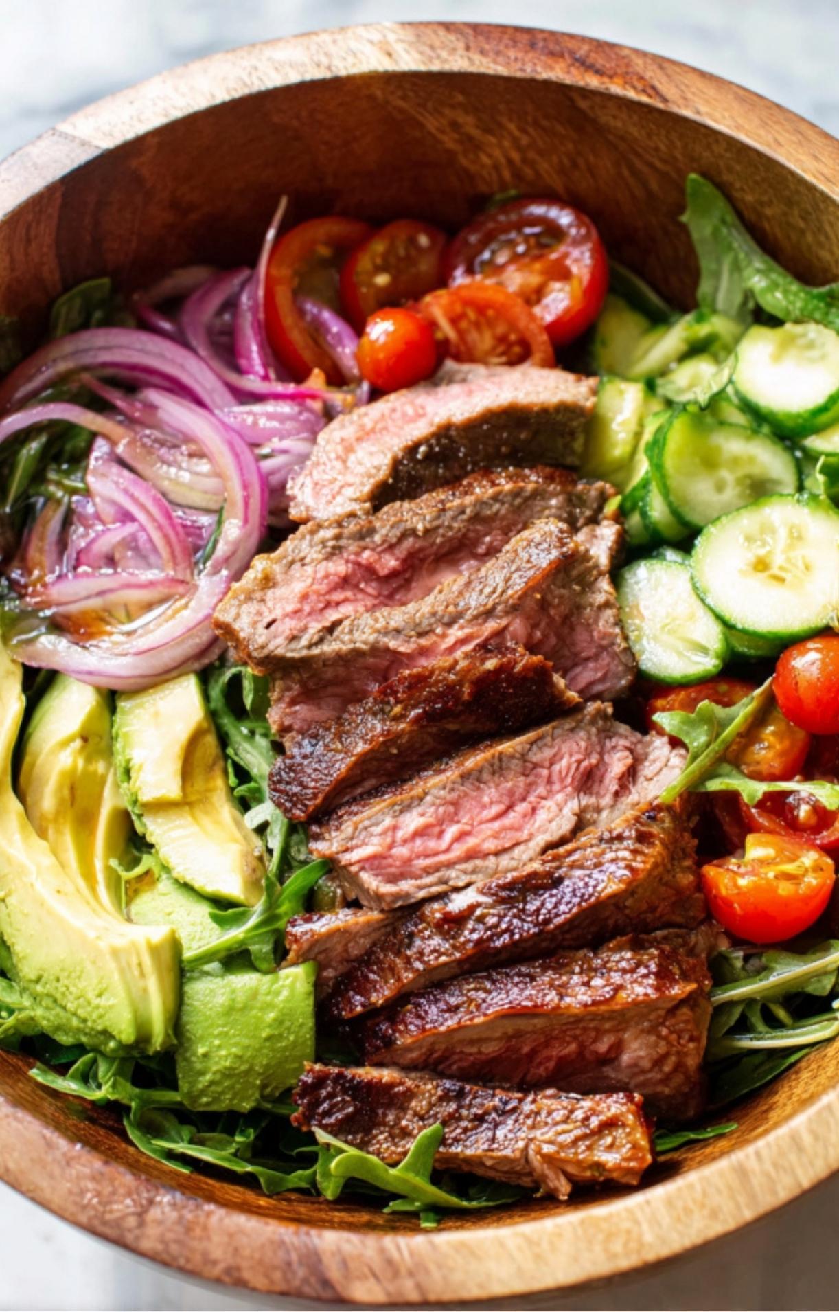 A wooden bowl organized with rows of sliced avocado, red onions, cherry tomatoes, cucumbers, and steak for a Steak Salad Recipe.