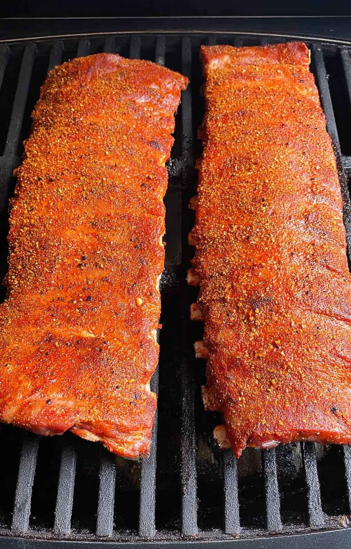 Two racks of seasoned baby back ribs smoking directly on the grates of a pellet grill.