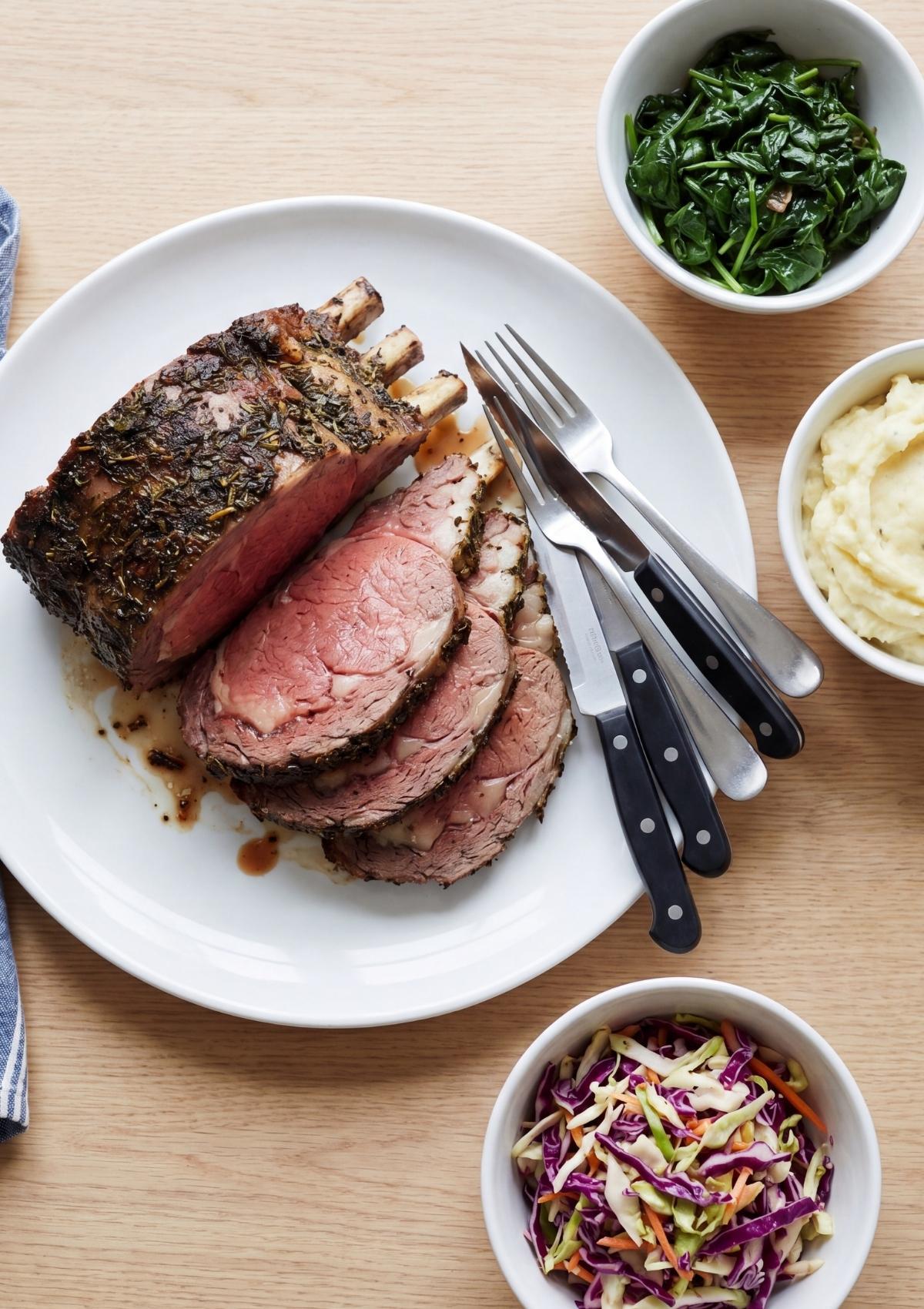 Slices of the standing rib roast served on a white plate with the bones attached, accompanied by bowls of mashed potatoes, spinach, and coleslaw.
