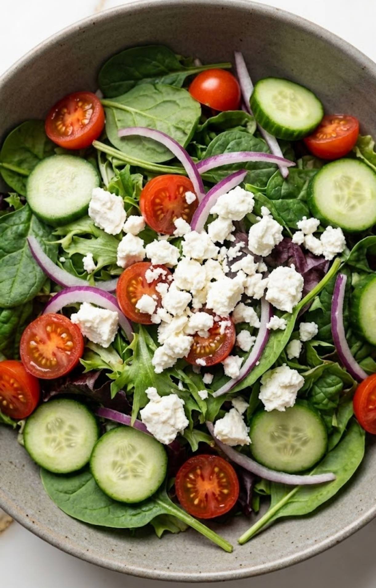A bowl filled with the base of a Healthy Steak Salad Recipe, including baby spinach, mixed greens, red onion slivers, cucumber rounds, halved tomatoes, and feta.