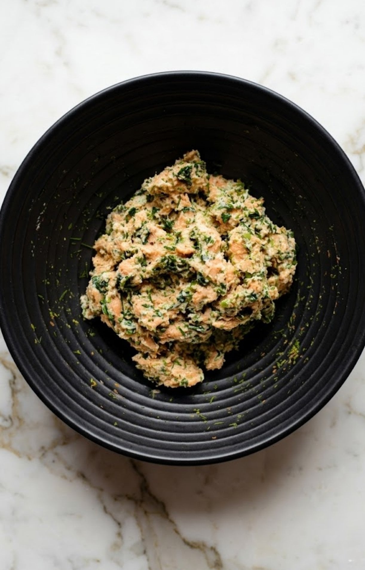 A top-down view of a black bowl containing a raw salmon burger mixture combined with fresh green herbs and breadcrumbs.