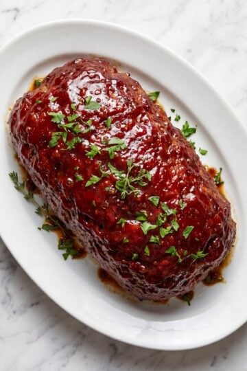 A whole cooked meatloaf with a shiny red glaze and fresh parsley garnish served on a white oval platter.