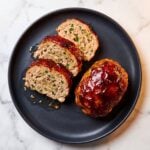 Slices of a juicy turkey meatloaf with a dark caramelized red glaze arranged on a black plate alongside a whole glazed mini meatloaf