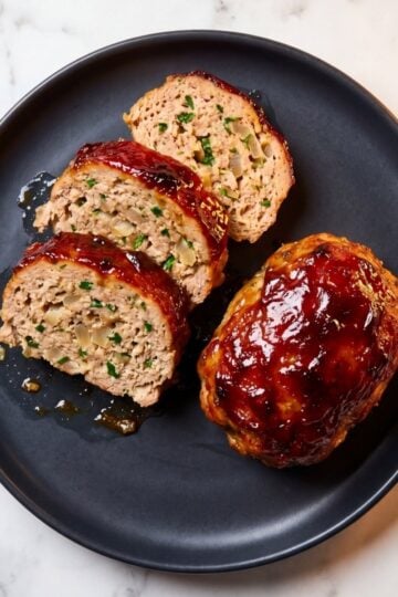 Slices of a juicy turkey meatloaf with a dark caramelized red glaze arranged on a black plate alongside a whole glazed mini meatloaf