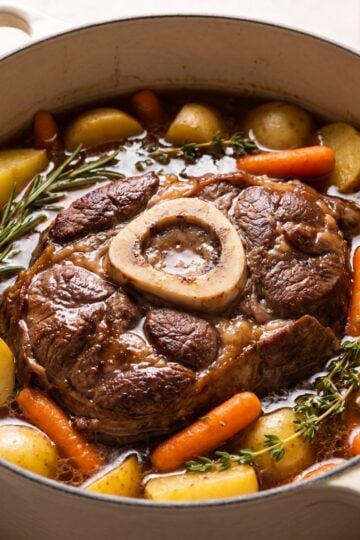 A bone-in beef arm roast pot roast slow-cooking in a rich broth with tender yellow potatoes and carrots inside a white Dutch oven.