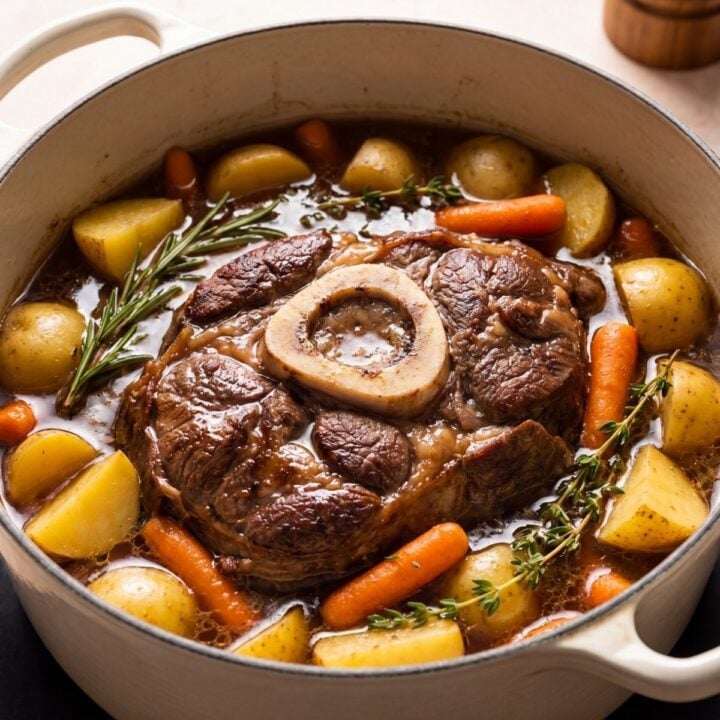 A bone-in beef arm roast pot roast slow-cooking in a rich broth with tender yellow potatoes and carrots inside a white Dutch oven.