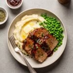 Two slices of juicy BBQ turkey meatloaf with a dark glaze served on a plate with mashed potatoes, a pat of melting butter, and green peas.