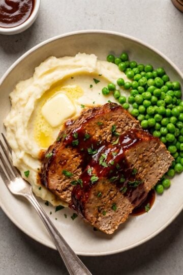 Two slices of juicy BBQ turkey meatloaf with a dark glaze served on a plate with mashed potatoes, a pat of melting butter, and green peas.