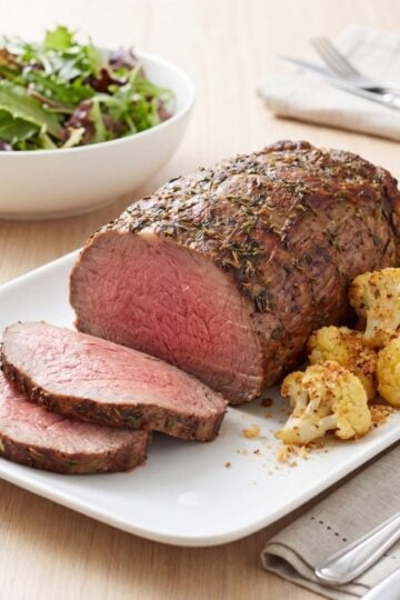 Sliced beef striploin roast served on a white platter with a side of crispy roasted cauliflower next to a bowl of green salad