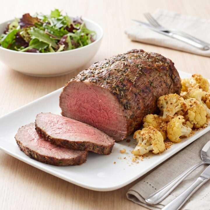 Sliced beef striploin roast served on a white platter with a side of crispy roasted cauliflower next to a bowl of green salad