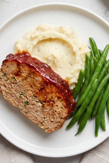 An overhead view of a thick slice of glazed turkey meatloaf served on a white plate with a scoop of mashed potatoes and a side of green beans.