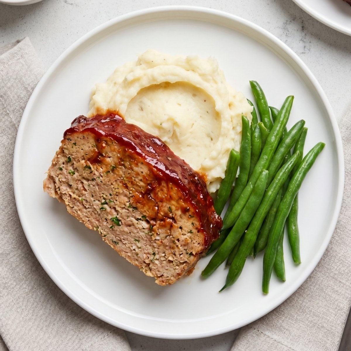 An overhead view of a thick slice of glazed turkey meatloaf served on a white plate with a scoop of mashed potatoes and a side of green beans.