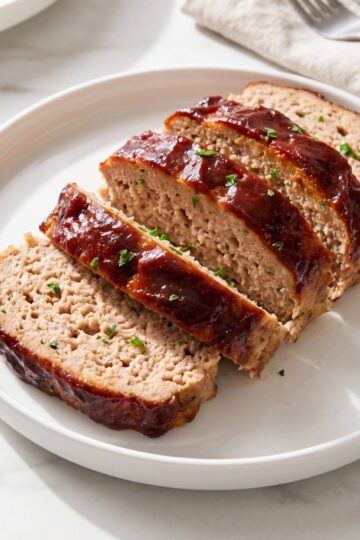 Sliced ground turkey meatloaf with a dark red glaze and fresh parsley garnish served on a white plate.