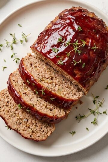 A sliced healthy turkey meatloaf with a dark red glaze and fresh thyme sprigs on a round white serving platter.