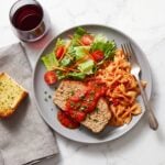 A grey plate holding a slice of turkey meatloaf topped with tomato sauce and fresh herbs, served alongside bow tie pasta, a side salad, garlic bread, and a glass of red wine