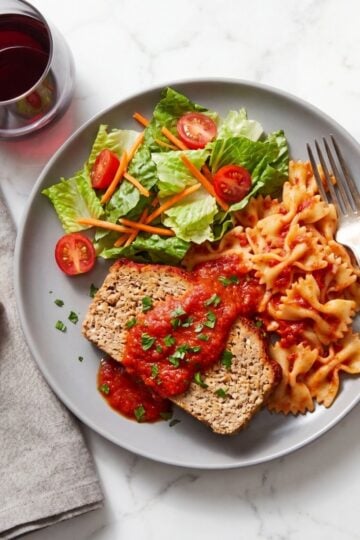 A grey plate holding a slice of turkey meatloaf topped with tomato sauce and fresh herbs, served alongside bow tie pasta, a side salad, garlic bread, and a glass of red wine