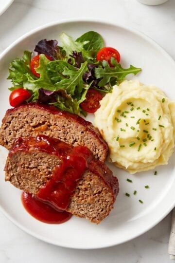 A close-up shot of two thick slices of meatloaf topped with a rich red glaze, paired with a swirl of creamy mashed potatoes garnished with chives and a side of mixed greens and cucumber.