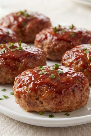 Six mini turkey meatloaves with a glossy red glaze arranged on a white serving platter and garnished with fresh chopped parsley