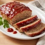 Slices of old fashioned turkey meatloaf with a thick red glaze served on a white platter alongside fresh parsley and dots of ketchup