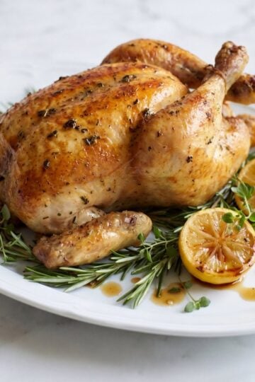 A whole golden brown roasted chicken resting on a bed of fresh rosemary and thyme with grilled lemon halves on a white serving plate