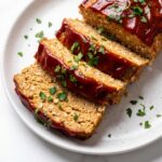 A close-up view of thick, tender slices of cooked turkey meatloaf arranged on a white plate, topped with a rich red glaze and sprinkled with chopped parsley.