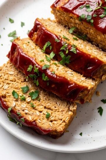 A close-up view of thick, tender slices of cooked turkey meatloaf arranged on a white plate, topped with a rich red glaze and sprinkled with chopped parsley.