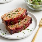 Two thick slices of juicy turkey and beef meatloaf topped with a rich red glaze and fresh parsley served on a white plate with a gold fork.