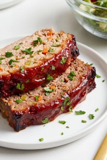 Two thick slices of juicy turkey and beef meatloaf topped with a rich red glaze and fresh parsley served on a white plate with a gold fork.