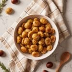 A white ceramic bowl filled with peeled roasted chestnuts coated in melted butter, cinnamon, and coarse sea salt, sitting on a plaid kitchen towel.