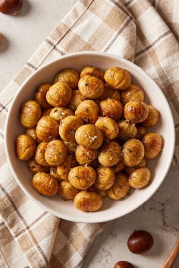 A white ceramic bowl filled with peeled roasted chestnuts coated in melted butter, cinnamon, and coarse sea salt, sitting on a plaid kitchen towel.