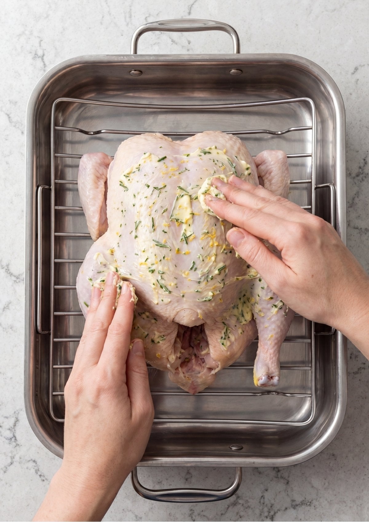 Overhead view of hands rubbing a textured herb and lemon zest butter mixture onto a raw whole chicken inside a stainless steel roasting pan