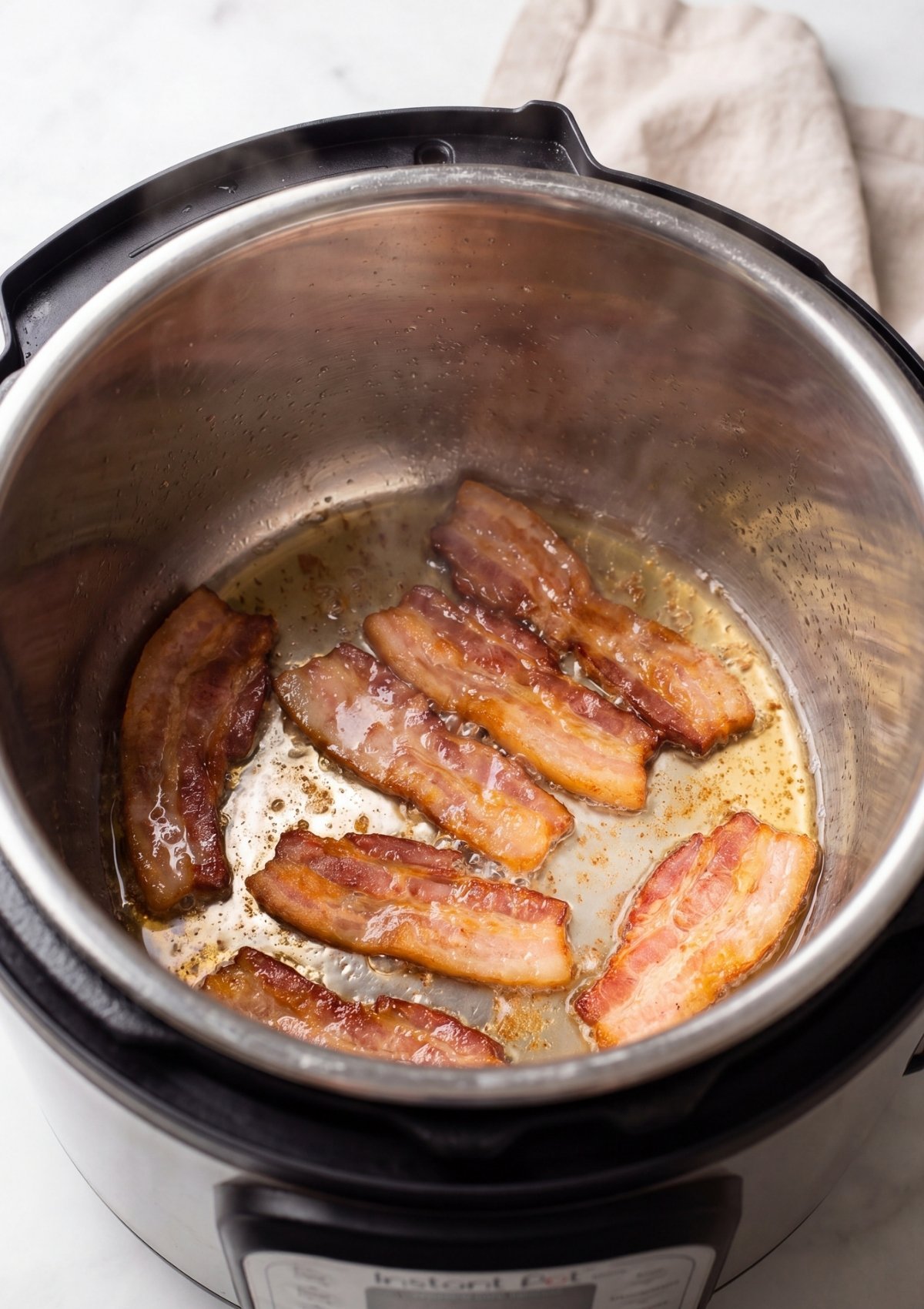 Sliced bacon strips sizzling and browning in the bottom of a stainless steel Instant Pot insert on a white marble countertop.