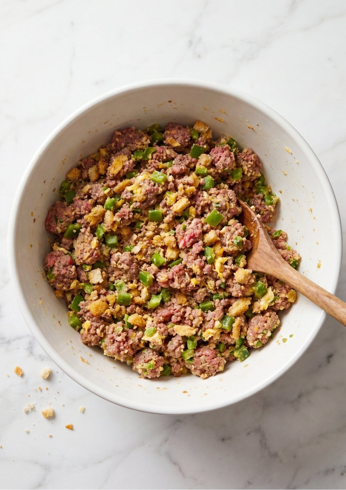 A white bowl filled with raw ground beef, breadcrumbs, green peppers, and egg being gently combined with a wooden spoon