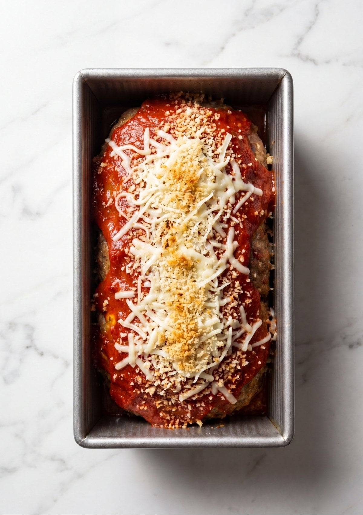 A raw meatloaf in a metal loaf pan topped with a layer of red sauce, shredded cheese, and golden breadcrumbs.
