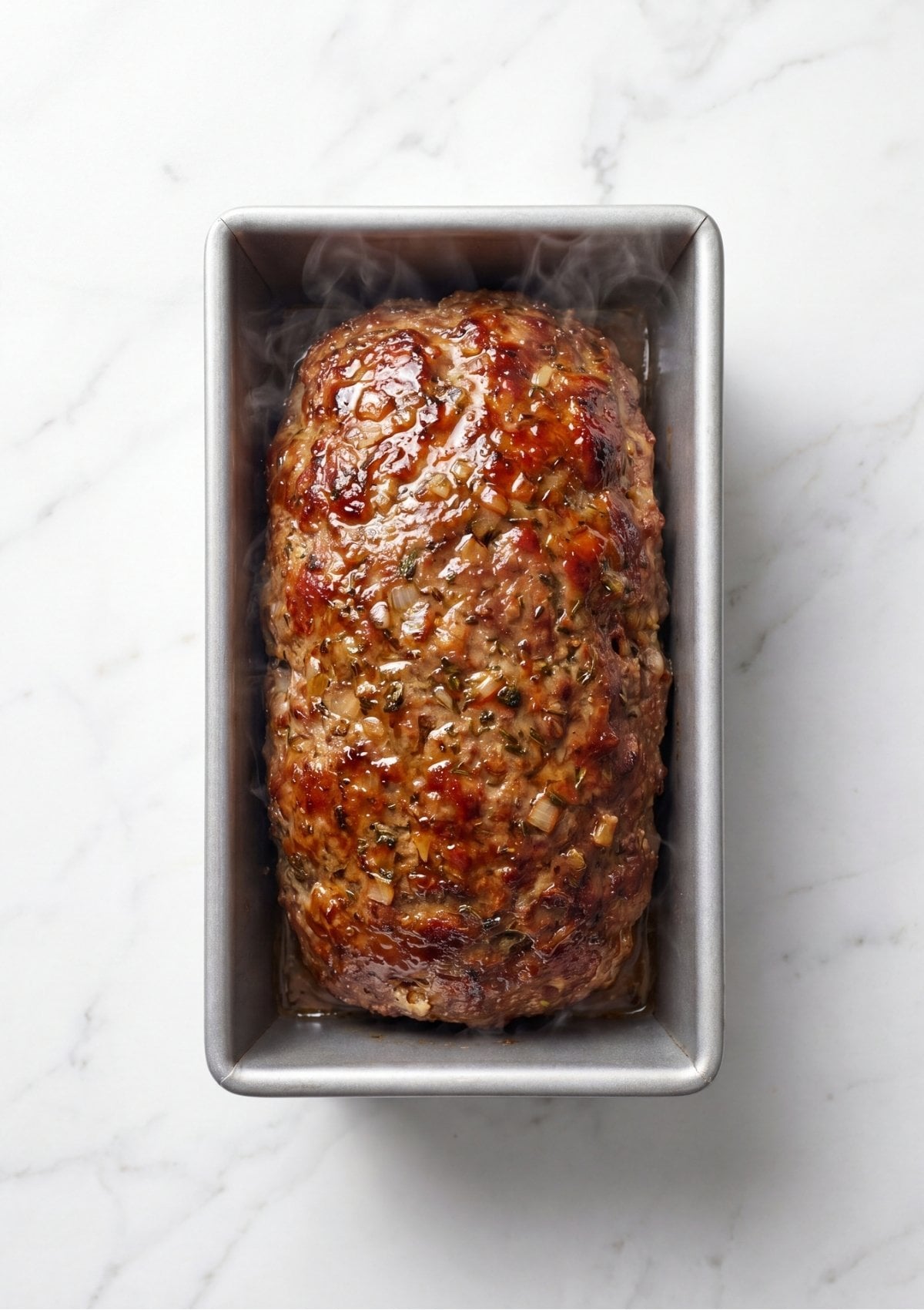 A top-down view of a steaming, fully cooked meatloaf inside a silver rectangular baking pan, showing a caramelized glaze and visible onions and herbs throughout the meat.
