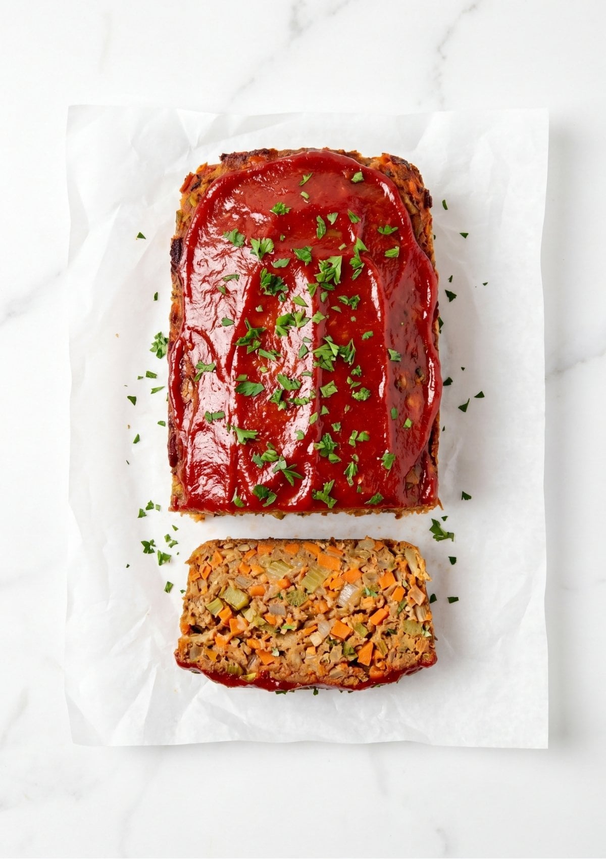 A sliced vegan meatloaf on white parchment paper showing a textured interior of chickpeas and diced vegetables with a red glaze and parsley garnish