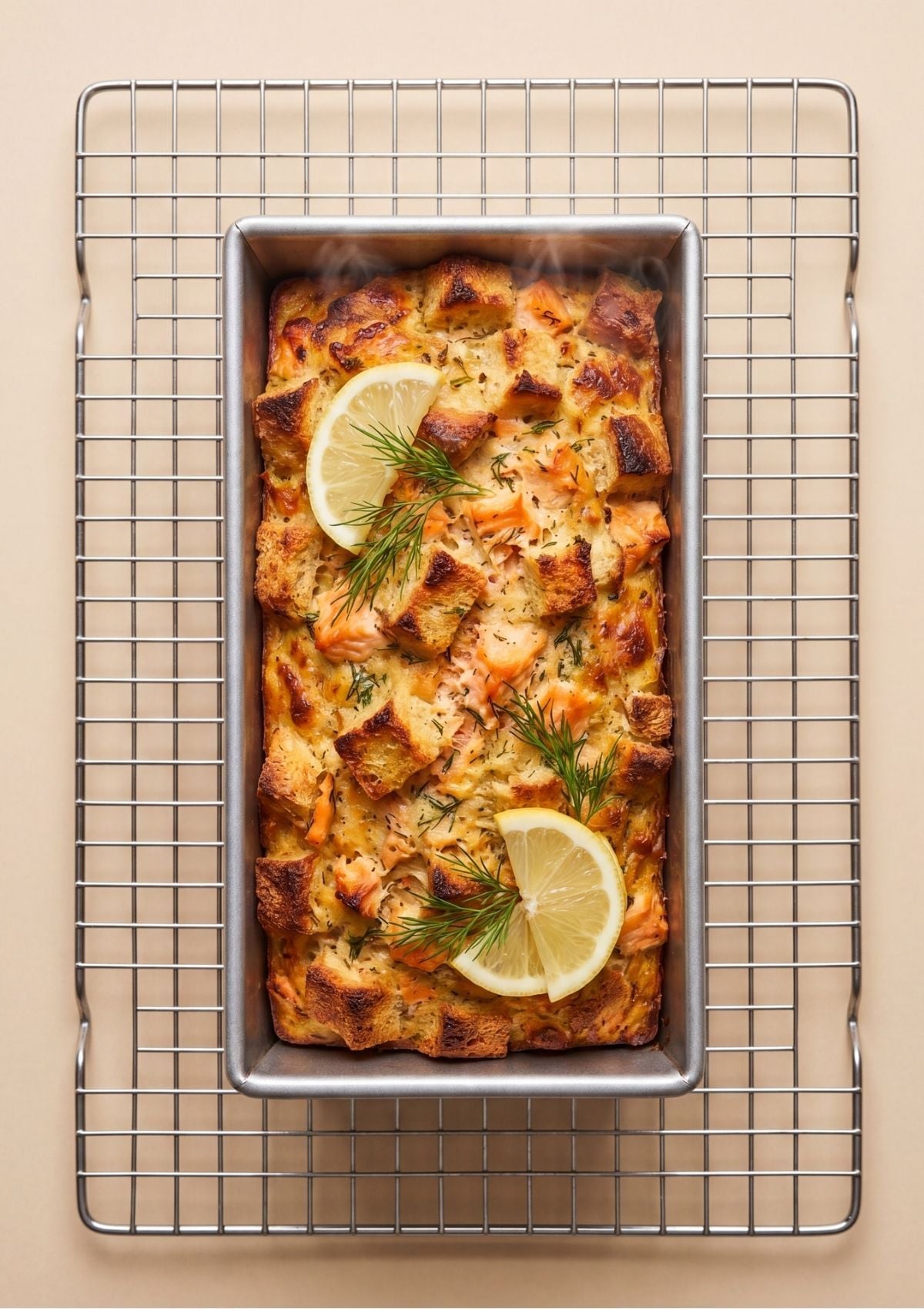 A freshly baked golden brown salmon loaf in a metal baking pan resting on a wire cooling rack garnished with lemon slices and fresh dill sprigs