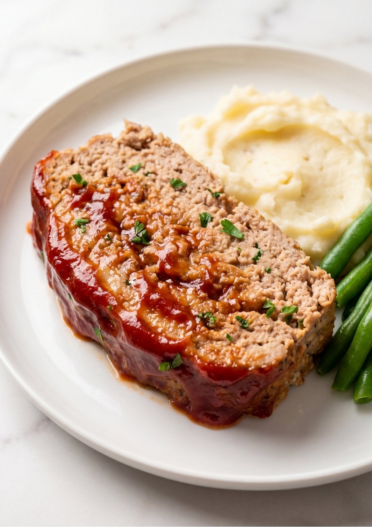 A thick slice of glazed turkey meatloaf served on a white plate with mashed potatoes and steamed green beans.